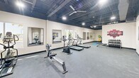 Located at 210 Reservation Way in Augusta, Georgia, this dedicated fitness facility offers a generously sized, single-floor exercise room designed to accommodate a comprehensive workout experience. The space features an open, industrial-style ceiling with exposed black steel framework, recessed lighting, and large ceiling fans that promote airflow and comfort throughout the room. The light-colored walls are complemented by dark base trim and durable gray carpet flooring, creating a clean, professional aesthetic throughout.

The exercise room is well-appointed with a diverse array of equipment, including treadmills, a stationary bike, an elliptical, a rowing machine, a cable machine, a weight bench, and free weights — providing options for both cardio and strength training. Multiple large wall-mounted mirrors are positioned strategically around the room, enhancing the sense of space and supporting proper form during exercise.

Natural light filters in through several windows along the perimeter walls, offering views of the surrounding outdoor greenery and adding to the bright, energizing atmosphere. Motivational artwork, including a large aerial stadium photograph, adorns the walls, lending the space a dynamic character. The layout is thoughtfully arranged to allow ample floor space for movement and functional training, making this fitness center a practical and inviting environment for users of all fitness levels.