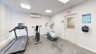 Located at 560 Birchmount Rd in Toronto, this dedicated exercise room offers a practical and well-equipped fitness space. The room features a drop-panel ceiling with recessed flat lighting, white walls, and a durable grey concrete-style floor, creating a clean and functional environment. Large vertical blinds line one wall, allowing for adjustable natural light throughout the space. A wall-mounted Split-system air conditioning unit ensures comfortable climate control during workouts. The cardio section includes a Spirit treadmill, an air assault bike, and an elliptical trainer, providing a variety of aerobic options. A multi-functional cable machine with a weight stack and bench caters to strength training needs, while a set of hex dumbbells on a rack and a BOSU balance trainer support free-weight and functional fitness exercises. An exposed brick column adds a subtle architectural character to the room. A bar and plate storage rack keeps accessories organized. The space is accessed through a door with a small window and is marked with a clearly visible exit sign, meeting safety standards. Overall, this exercise room is efficiently laid out to support a comprehensive fitness routine.