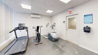 Located at 560 Birchmount Rd in Toronto, this dedicated exercise room offers a practical and well-equipped fitness space. The room features a drop-panel ceiling with recessed flat lighting, white walls, and a durable grey concrete-style floor, creating a clean and functional environment. Large vertical blinds line one wall, allowing for adjustable natural light throughout the space. A wall-mounted Split-system air conditioning unit ensures comfortable climate control during workouts. The cardio section includes a Spirit treadmill, an air assault bike, and an elliptical trainer, providing a variety of aerobic options. A multi-functional cable machine with a weight stack and bench caters to strength training needs, while a set of hex dumbbells on a rack and a BOSU balance trainer support free-weight and functional fitness exercises. An exposed brick column adds a subtle architectural character to the room. A bar and plate storage rack keeps accessories organized. The space is accessed through a door with a small window and is marked with a clearly visible exit sign, meeting safety standards. Overall, this exercise room is efficiently laid out to support a comprehensive fitness routine.