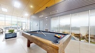 Located at 1240 Benchmark Park Dr in Columbus, Ohio, this single-floor community amenity space offers a well-designed, open-plan layout that blends recreation and relaxation. The interior is defined by soaring double-height ceilings clad in warm, paneled finishes on one side and horizontal grey siding on the other, creating a striking visual contrast throughout the space.

Natural light floods the interior through expansive floor-to-ceiling glass walls and large skylights, connecting the indoors with views of the surrounding outdoors. Polished concrete flooring runs throughout, lending the space a clean, contemporary feel.

A centerpiece rustic-wood billiards table anchors the recreational zone, complemented by bar-height seating and a counter area ideal for socializing. A comfortable lounge area nearby features a plush grey sofa, a flat-screen TV mounted on the paneled wall, and curated greenery that adds warmth to the modern aesthetic.

A casual dining nook with a solid-wood table and leather chairs sits adjacent to frosted-glass partitioned rooms, which appear to serve as private offices or meeting spaces accessible from the main floor. The layout flows seamlessly from one zone to the next, making this a versatile and inviting communal gathering destination.