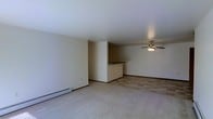 This single-floor space offers a straightforward and functional layout with a clean, neutral aesthetic throughout. The main living area is generously proportioned, featuring wall-to-wall carpeting that adds warmth underfoot. A ceiling fan with a built-in light fixture is centrally positioned, providing both air circulation and ambient lighting to the room.

A defining architectural detail is the partial wall peninsula that separates the living area from the kitchen space beyond. This half-wall partition creates a natural division between the two areas while maintaining an open, connected feel. The kitchen area, visible beyond the peninsula, features tile or vinyl flooring and a dark-topped counter surface along the partition, offering a convenient bar-style ledge.

The walls throughout are finished in a soft, neutral tone, complementing the light-colored carpet and contributing to the overall bright ambiance of the space. Baseboard heating units are present along the exterior wall, indicating a practical heating solution integrated into the room. Dark base trim runs along the perimeter, providing a subtle contrast to the light walls and flooring. The overall design is simple and utilitarian, making the space versatile and easy to furnish.