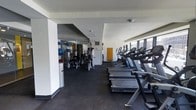 This fitness center is a comprehensively outfitted exercise facility designed to meet a wide range of workout needs. The space is organized into distinct functional zones, allowing users to move seamlessly between cardiovascular training and strength work. Along one side of the room, a row of Cybex treadmills and elliptical machines lines the perimeter, positioned beside large windows that invite natural light and provide open views to the outside — creating an energizing environment for cardio workouts. The flooring throughout is a durable dark rubber mat, characteristic of professional-grade fitness facilities and ideal for both equipment support and user safety. On the opposite side, a well-stocked strength training area houses a variety of cable machines, resistance equipment, and free weights, supported by mirrored walls that enhance the sense of space and allow users to monitor their form. The ceiling is clean and white with recessed lighting supplementing the natural light from the windows. The overall aesthetic is modern and functional, with a neutral white and grey color palette that keeps the environment feeling open and focused. The single-floor layout ensures easy accessibility throughout, making this a practical and well-designed space for a full fitness experience.