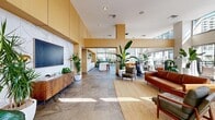Located in the heart of downtown Los Angeles at 705 W 9th St, this expansive single-floor amenity lounge offers a generously proportioned, open-plan space designed for both relaxation and social engagement. The architecture is defined by soaring double-height ceilings and an abundance of floor-to-ceiling glazing that floods the interior with natural light while framing striking views of the surrounding cityscape. Large-format stone tile flooring runs throughout, grounding the space with a clean, contemporary aesthetic.

The lounge area features multiple thoughtfully arranged seating vignettes, including rich cognac leather sofas, a bold velvet sectional in warm amber tones, and sculptural accent chairs, all anchored by layered area rugs that define each zone. Lush tropical plants are positioned throughout, adding vibrancy and a sense of organic warmth to the modern interior.

A full-length kitchen runs along one wall, highlighted by warm wood cabinetry, a marble-slab backsplash, open display shelving, and a substantial white island with bar seating — ideal for communal gatherings. A wall-mounted display screen and textured accent panels complete the entertainment area nearby.

Sliding glass doors connect the interior directly to an outdoor terrace, where additional seating is available against the backdrop of the downtown Los Angeles skyline, creating a seamless indoor-outdoor experience.