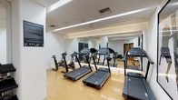 This fitness center offers a comprehensive and thoughtfully designed workout environment within a single-floor layout. The space features clean white walls, warm cork-style flooring, and recessed linear LED lighting that creates a bright, energizing atmosphere throughout.

The cardio zone is prominently appointed with multiple treadmills and stationary cycling bikes arranged in an orderly row, allowing users ample space to exercise comfortably. Floor-to-ceiling mirrors line key sections of the walls, visually expanding the space and allowing users to monitor their form during workouts.

The strength training area includes a well-stocked dumbbell rack with a wide range of hex weights displayed on a multi-tiered stand, accompanied by an adjustable workout bench. A cable resistance machine is also available, catering to a variety of strength exercises. A wall-mounted flat-screen television provides entertainment throughout the space.

Near the entrance, a wooden double door provides access, flanked by a sleek wooden bench with a lower storage shelf and a waste bin — offering a convenient area to prepare before or cool down after a workout. Fitness center rules are clearly posted, ensuring a safe and organized environment for all users. The overall design is modern and functional, prioritizing cleanliness, accessibility, and a motivating atmosphere.
