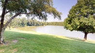 This single-level outdoor space presents a beautifully natural lakefront setting, characterized by its open, expansive grounds and calming waterside atmosphere. The centerpiece of the space is a generous body of water, its calm surface reflecting the surrounding landscape and open sky, creating a mirror-like visual effect that adds depth and serenity to the environment. A well-maintained, lush green lawn slopes gently toward the water's edge, providing an inviting and accessible transition between land and lake. Mature deciduous trees frame the scene, their broad canopies extending overhead to offer natural shade and a sense of established, enduring beauty. The tree cover along the far shoreline creates a dense green backdrop that encloses the space, lending it a private and secluded character. The overall ambiance is one of quiet natural beauty — ideal for outdoor leisure, reflection, or recreation. The open sky above and the interplay of sunlight across the water and grass contribute to a bright, airy atmosphere throughout the day. This outdoor space offers a rare combination of open lawn, water frontage, and mature tree coverage that together create a distinctly peaceful and picturesque natural environment.