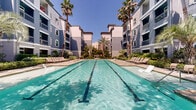 Located at 11070 Katy Fwy in Houston, TX, this outdoor amenity space is the centerpiece of a well-appointed multi-story residential community. The architecture features a distinctive blue-gray exterior with arched windows, decorative shutters, and private balconies, creating an elegant Mediterranean-inspired aesthetic that frames the courtyard on multiple sides.

At the heart of the space is a generously sized lap pool with crystal-clear turquoise water, accented by stone tile coping and surrounded by a broad pool deck fitted with lounge chairs — ideal for relaxing under the Texas sun. Tall palm trees line the pool area, lending a resort-like ambiance that elevates the outdoor experience.

The grounds are thoughtfully landscaped with a rich variety of mature trees, tropical plants, manicured shrubs, and ground cover that create a lush, verdant setting. Stone-paved pathways wind through the greenery, connecting different areas of the courtyard and leading to gated pool entrances framed by canopy trees.

Additional green lawn areas are interspersed throughout the complex, offering open, park-like spaces. The overall design balances openness with privacy, creating an inviting retreat within an urban setting. The space is well-maintained and reflects a strong commitment to outdoor living and community amenity.