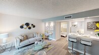 This single-floor residence offers a thoughtfully designed open-concept layout that maximizes functionality and flow throughout the space. The living area is bright and welcoming, featuring light hardwood-style flooring, a large window with vertical blinds that allows natural light to fill the room, and a textured ceiling. A dedicated nook adjacent to the living area provides a compact workspace, illuminated by recessed lighting and framed by a soft blue accent wall that carries through much of the interior, lending a cohesive, calming palette to the home.

The kitchen is efficiently laid out with gray shaker-style cabinetry, white subway tile backsplash, and a light quartz countertop that extends into a breakfast bar with bar seating — ideal for casual dining. A white refrigerator and ample counter space complete the functional kitchen.

The bedroom is a comfortable retreat, highlighted by the same blue accent wall, a built-in open closet with shelving and hanging space, and a window with vertical blinds providing natural light and privacy. Light hardwood-style flooring continues throughout.

The bathroom is neatly appointed with a white vanity, a shower-tub combination with a decorative curtain, and brushed nickel fixtures, offering a clean and practical design.