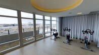 This single-floor fitness space is designed with both functionality and aesthetics in mind. The room is anchored by a striking circular recessed ceiling detail, illuminated with warm yellow ambient lighting that adds a distinctive architectural focal point overhead. The light hardwood flooring complements the neutral grey tones throughout, creating a clean and contemporary atmosphere.

Floor-to-ceiling windows wrap along one curved wall, flooding the space with abundant natural light while framing expansive views of the surrounding cityscape and landscape beyond. The elevated vantage point enhances the open, energizing feel of the room. On the opposite side, floor-length grey curtains offer the option for privacy when desired, adding a soft textural contrast to the otherwise sleek interior.

The fitness area is equipped with stationary cycling bikes, thoughtfully positioned to take advantage of the panoramic views during workouts. The curved layout of the room contributes to a sense of fluid, uninterrupted space. Overall, the combination of architectural detail, generous natural light, and well-placed equipment makes this a bright, motivating, and well-appointed fitness environment.