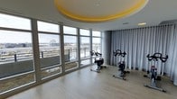 This single-floor fitness space is designed with both functionality and aesthetics in mind. The room is anchored by a striking circular recessed ceiling detail, illuminated with warm yellow ambient lighting that adds a distinctive architectural focal point overhead. The light hardwood flooring complements the neutral grey tones throughout, creating a clean and contemporary atmosphere.

Floor-to-ceiling windows wrap along one curved wall, flooding the space with abundant natural light while framing expansive views of the surrounding cityscape and landscape beyond. The elevated vantage point enhances the open, energizing feel of the room. On the opposite side, floor-length grey curtains offer the option for privacy when desired, adding a soft textural contrast to the otherwise sleek interior.

The fitness area is equipped with stationary cycling bikes, thoughtfully positioned to take advantage of the panoramic views during workouts. The curved layout of the room contributes to a sense of fluid, uninterrupted space. Overall, the combination of architectural detail, generous natural light, and well-placed equipment makes this a bright, motivating, and well-appointed fitness environment.