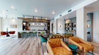 This stylish single-floor communal space is designed with an open-concept layout that seamlessly integrates multiple functional zones. Light hardwood flooring runs throughout, lending warmth and continuity to the entire space.

The kitchen area is a standout feature, anchored by a large marble-topped island with vibrant green bar stools. Dark cabinetry with open shelving, a textured tile backsplash, and a striking wooden ceiling beam overhead create a bold, contemporary aesthetic. Sculptural globe pendant lights add an artistic focal point above the island.

Adjacent to the kitchen, a generously sized dining and gathering area offers a dark communal table surrounded by teal velvet chairs, complemented by built-in teal shelving units stocked with books and décor. A media lounge area features an olive-green accent wall with paneling, a wall-mounted television, and eclectic seating arrangements including tufted leather chairs and colorful accent pieces.

Toward the entrance, floor-to-ceiling glass partitions and large double glass doors open to an outdoor terrace, flooding the space with natural light and offering views of the surrounding exterior. A cozy seating nook with jewel-toned armchairs and vibrant patterned cushions adds an inviting, relaxed corner. Overall, the space presents a thoughtfully curated, modern design with a lively and welcoming ambiance.