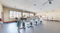 This well-appointed fitness center at Harrison Creek in Petersburg, Virginia, offers a generously sized, single-floor workout facility designed to support a full range of exercise routines. The open floor plan provides ample space between equipment stations, allowing for comfortable movement throughout the room.

The facility is outfitted with an extensive array of Legend-brand strength machines, cable systems, and a dedicated free weights area featuring a full dumbbell rack. Cardio equipment includes multiple treadmills, an elliptical trainer, a stationary bike, and a rowing machine, catering to a variety of fitness preferences. A squat rack and weight bench further support strength training needs.

Large, framed windows line the walls, flooding the space with natural light and offering views of the surrounding community. Ceiling fans are distributed throughout the room, complementing the recessed lighting and HVAC system to maintain a comfortable workout environment. Durable rubber flooring covers the entire space, providing safety and functionality.

Full-length mirrors along one wall visually expand the room while also serving a practical purpose for form checking. A scenic coastal mural adorns the upper wall, adding a decorative touch to the otherwise functional space. Wall-mounted televisions provide additional entertainment during workouts. The facility is accessible via wood-trimmed glass doors, consistent with the community's overall aesthetic.