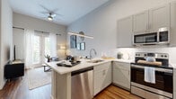 Located at 1610 Hill St in Grand Prairie, TX, this well-designed one-bedroom, one-bathroom residence offers a thoughtful layout across a single floor. The open-concept kitchen and living area is immediately impressive, anchored by soaring vaulted ceilings and a sleek peninsula with bar seating and a pendant light fixture. The kitchen is outfitted with gray shaker cabinetry, white quartz countertops, a subway tile backsplash, and a full suite of stainless steel appliances including a refrigerator, range, built-in microwave, and dishwasher. Rich wood-look flooring runs throughout the main living areas, complementing the bright, neutral palette. The living room benefits from natural light streaming through French doors, which open to the outdoors and are framed by floor-to-ceiling curtains. A ceiling fan adds comfort overhead. The bedroom features plush carpeting, a large window with outdoor views, high ceilings, and a dedicated walk-in closet with built-in shelving and hanging rods. The bathroom is finished with a gray vanity, white quartz countertop, large mirror, pendant lighting, and a full tub-shower combination with tile surround and wood-look flooring throughout.