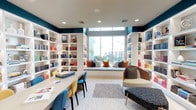 This thoughtfully designed library offers a refined blend of functionality and comfort across a single level. The space is anchored by expansive floor-to-ceiling white built-in shelving units that line the walls, filled with a curated collection of books and decorative objects. Bold teal accent walls provide a striking contrast against the bright white shelving and light flooring, creating a visually dynamic yet cohesive aesthetic.

A standout feature is the built-in window seat nestled between flanking bookshelves, fitted with a plush cushion and an array of colorful throw pillows in warm orange, navy, and neutral tones. The oversized window above the seat frames outdoor views and floods the nook with natural light, making it an ideal reading retreat.

A long communal work table with colorful modern chairs in blue, yellow, and teal serves as a central gathering point for collaborative study or reading. Adjacent lounge seating with contemporary chairs and an ottoman offers a more relaxed setting.

A glass-walled enclosure connects seamlessly to a separate workspace equipped with desks and ergonomic seating, maintaining an open and transparent feel. Recessed ceiling lighting throughout ensures the entire space remains well-illuminated and inviting.