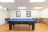 Located at 1431 Lakeshore Rd in Burlington, Ontario, this single-floor amenity room serves as a versatile recreational and social gathering space. The room is finished with warm hardwood-style flooring throughout the main area, transitioning to tile near the kitchenette, and features a suspended drop ceiling equipped with large skylight-style flush light panels that flood the interior with bright, even illumination. A row of clerestory windows along one wall allows additional natural light to filter in.

The space is thoughtfully organized into distinct activity zones. A full-size pool table with a vivid blue felt surface anchors the recreational area, complemented by a shuffleboard table along one wall and a round game table with chairs for additional tabletop activities. Lounge seating, including a large dark leather sofa and a pair of patterned armchairs with glass-topped coffee tables, creates a comfortable gathering area. A compact kitchenette with a white countertop, stove, refrigerator, and range hood provides functional support for events and gatherings. A small bookshelf stocked with reading materials adds a quiet corner for those seeking a more relaxed activity. Framed landscape artwork decorates the cream-coloured walls throughout, contributing to the warm and welcoming ambiance of the space.