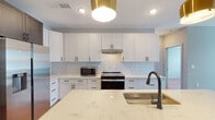 This thoughtfully designed single-floor residence blends modern aesthetics with functional living. At its heart is an open-concept kitchen that serves as the focal point of the space. Crisp white shaker cabinetry with matte black hardware lines the walls, complemented by a classic white subway tile backsplash. A generous central island topped with veined quartz countertops provides ample workspace and includes an undermount sink with a matte black faucet, illuminated by a pair of striking brushed gold pendant lights. Stainless steel appliances, including a French door refrigerator and a slide-in range, round out the well-appointed kitchen. The space flows seamlessly into the living area, where warm-toned hardwood flooring extends throughout. Bedrooms are bright and airy, each featuring ceiling fans and large windows that invite natural light and offer views of the surrounding neighborhood. The bathroom is spacious and accessible, outfitted with large-format dark tile flooring, a dual-sink vanity with gray quartz countertops, white cabinetry, and matte black fixtures. Accessibility grab bars reflect a commitment to inclusive design. The overall ambiance is clean, contemporary, and move-in ready.