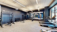 Located at 30 W Broad St in Bethlehem, PA, this contemporary fitness center offers a thoughtfully designed exercise space across a single floor. The main workout area is characterized by deep navy accent walls, a neutral carpet tile floor, and a suspended linear lighting system set within a clean drop-tile ceiling, creating a focused and energizing atmosphere. Floor-to-ceiling mirror panels line one wall, visually expanding the space and supporting form-check during workouts. The cardio zone features a row of Spirit treadmills, elliptical trainers, and a stair climber positioned in front of expansive floor-to-ceiling windows that flood the area with natural light and provide open views of the surrounding streetscape. On the strength side, the room is outfitted with a multi-station cable machine, a Smith machine loaded with weight plates, a full dumbbell rack with a flat bench, and additional weight training equipment. A separate enclosed studio room, accessed through sleek black-framed glass doors, provides a dedicated space for functional training and flexibility work, equipped with kettlebells, resistance bands, stability balls, yoga mats, and a wall-mounted display screen. A decorative accent wall with an abstract brushstroke mural adds an artistic touch to this area. Storage cabinetry and accessible HVAC vents complete the practical amenities of this well-appointed fitness facility.