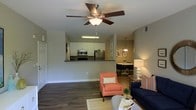This single-story space showcases an open-concept floor plan that integrates the living, kitchen, and dining areas into one cohesive layout. The living area is anchored by durable wood-style flooring and features a ceiling fan with a built-in light fixture, providing both comfort and illumination. A neutral color palette on the walls creates a clean, versatile backdrop throughout the space.

The kitchen is positioned behind a raised peninsula counter, which serves as both a functional workspace and a visual divider between the living and cooking areas. White cabinetry lines the kitchen walls, complemented by stainless steel appliances, including a refrigerator and microwave, adding a modern touch. Under-cabinet lighting enhances visibility and ambiance.

Adjacent to the kitchen, the dining area is defined by a stylish pendant light hanging above the table, creating an intimate setting for meals. The open sightlines between all three areas promote a sense of spaciousness and connectivity, making the layout well-suited for both daily living and entertaining. Overall, the space offers a functional and cohesive design with consistent finishes throughout.