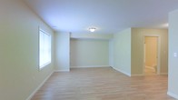 This single-floor space offers a clean and inviting atmosphere, thoughtfully designed with simplicity and functionality in mind. The open main living area immediately draws attention with its smooth light hardwood flooring, which runs throughout and lends a warm, airy feel to the interior. Walls finished in a soft, pale yellow-green hue create a calm and neutral backdrop, complementing the bright white ceiling above. Natural light streams in through a large window, enhancing the sense of openness and making the space feel welcoming throughout the day. A flush-mount ceiling light fixture provides additional illumination, ensuring the room remains well-lit regardless of the time of day. White baseboards provide a crisp contrast against the light walls and flooring, contributing to the overall tidy and polished aesthetic. A doorway visible toward the rear of the main area hints at additional rooms or storage beyond, offering practical utility. The layout is straightforward and versatile, providing ample room to configure furnishings to suit a variety of needs. The overall ambiance is bright, clean, and move-in ready, making it an ideal canvas for a range of uses and interior styles.