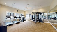 This dedicated fitness facility is thoughtfully designed across two interconnected exercise rooms, offering a comprehensive workout environment. The main exercise room is bright and spacious, bathed in natural light from large multi-pane windows along the walls. The room features neutral beige carpet tile flooring, warm tan walls, and a ceiling fan complemented by recessed lighting overhead. Cardio equipment, including treadmills and an elliptical trainer, lines one side of the room, while a multi-station cable machine and a punching bag occupy another zone. Floor-to-ceiling mirrors along one wall visually expand the space and support form-checking during workouts. The second exercise room is equally well-appointed, housing stationary bikes, a recumbent bike, a dumbbell rack with a weight bench, and stability balls, offering a versatile free weights and cycling area. A ceiling fan and recessed lighting keep the space comfortable and well-lit. An adjacent smaller room, accessible through a doorway, features a wall-mounted television and storage, serving as a flexible multipurpose space. Motivational fitness posters and a wall clock round out the practical, performance-focused atmosphere throughout.