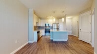 Located at 5671 Mauna Loa Blvd in Sarasota, Florida, this single-floor residence offers a thoughtfully designed open-concept layout with clean, contemporary finishes throughout. The kitchen serves as the heart of the home, showcasing crisp white cabinetry with glass-front upper panels, a tasteful tile backsplash, gleaming stainless steel appliances including a range, microwave, refrigerator, and dishwasher, and a spacious center island with a light quartz countertop and built-in shelving. Elegant pendant lighting and a track light fixture illuminate the space, complementing the warm wood-look plank flooring that flows seamlessly through the main living areas. The open layout connects the kitchen to a generous living area, which benefits from natural light streaming through slatted blinds and is topped with a ceiling fan for comfort. The bedroom features plush carpeting, a ceiling fan, and a double window that invites natural light. The bathroom is finished with neutral tile flooring, a white vanity with multiple drawers, and clean, simple fixtures. A dedicated laundry area houses a full-size side-by-side washer and dryer with overhead wire shelving. The overall ambiance is bright, airy, and move-in ready, reflecting a modern Florida lifestyle.