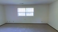 This single-floor space offers a clean, functional layout with a simple and understated aesthetic. The main room is generously proportioned, featuring wall-to-wall carpeting in a neutral tone that contributes to a quiet, comfortable atmosphere. Smooth, white walls provide a bright and versatile backdrop, making the space well-suited for a variety of uses and furnishing styles. A wide, double-panel window fitted with horizontal blinds serves as a focal point along one wall, allowing an abundance of natural light to flow into the room and creating a connection to the outdoors. The ceiling is smooth and finished with a centrally placed ventilation grille, indicating a built-in climate control system. Baseboards and electrical outlets are neatly integrated throughout, reflecting a straightforward and practical interior finish. The overall ambiance is open, airy, and move-in ready, with a neutral palette that provides a blank canvas for personalization. The single-floor layout ensures ease of accessibility and navigation throughout the space.