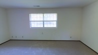 This single-floor space offers a clean, functional layout with a simple and understated aesthetic. The main room is generously proportioned, featuring wall-to-wall carpeting in a neutral tone that contributes to a quiet, comfortable atmosphere. Smooth, white walls provide a bright and versatile backdrop, making the space well-suited for a variety of uses and furnishing styles. A wide, double-panel window fitted with horizontal blinds serves as a focal point along one wall, allowing an abundance of natural light to flow into the room and creating a connection to the outdoors. The ceiling is smooth and finished with a centrally placed ventilation grille, indicating a built-in climate control system. Baseboards and electrical outlets are neatly integrated throughout, reflecting a straightforward and practical interior finish. The overall ambiance is open, airy, and move-in ready, with a neutral palette that provides a blank canvas for personalization. The single-floor layout ensures ease of accessibility and navigation throughout the space.