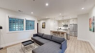 Located at 225 Vine St NW in Washington, DC's Shepherd Park neighborhood, this thoughtfully designed single-floor residence showcases a clean, modern aesthetic throughout. The open-concept living and kitchen area serves as the heart of the space, where light wood-tone flooring, recessed lighting, and crisp white walls create a bright and inviting atmosphere. The kitchen is well-appointed with white shaker-style cabinetry extending to the ceiling, a classic white tile backsplash, stainless steel appliances including a refrigerator, range, and microwave, and a peninsula with bar seating finished with warm wood stools — ideal for casual dining or entertaining. The living area is generously proportioned, accommodating a large sectional sofa, and features glass block windows that invite natural light while maintaining privacy. A wall-mounted television and recessed lighting make it a comfortable gathering space. The bedrooms are serene and functional, each with light wood flooring, clean white walls, and notable dark-framed double doors providing ample closet storage. The bathrooms are finished with large-format light tile flooring, black matte fixtures, and a glass-enclosed shower, reflecting a consistent modern design language. Dark espresso-toned door frames appear throughout, offering a striking contrast to the otherwise neutral palette and lending the space a refined, cohesive character.