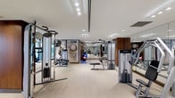 This modern fitness center offers a comprehensive workout environment designed to accommodate a full range of exercise needs. The space is generously proportioned, with a clean, open layout that allows for easy movement between equipment stations. The ceiling is bright and well-lit, featuring recessed lighting that evenly illuminates the entire floor, creating an energizing atmosphere conducive to physical activity. The flooring is finished in a neutral, durable material suited for high-traffic fitness use. A broad selection of professional-grade strength and resistance machines lines the space, including cable systems and multi-function units from recognized brands such as Life Fitness. Free weights, medicine balls, and stability balls are also present, catering to varied training styles. A standout decorative feature is a large-scale vintage U.S. postage stamp wall graphic, adding a unique artistic element to the otherwise sleek interior. Full-length mirrors are strategically placed to support proper form during workouts and to visually expand the space. Warm wood accent panels provide a refined contrast to the predominantly white and grey palette. The overall ambiance is clean, professional, and motivating, reflecting a high-end fitness facility well-suited for both casual and dedicated training.