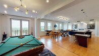 This expansive single-floor space blends refined architectural detailing with contemporary furnishings across several well-defined living and entertainment areas. Throughout, warm hardwood flooring, crown molding, recessed lighting, and decorative column accents establish a cohesive and polished aesthetic.

The kitchen is a standout, featuring sleek white cabinetry, a subway-tile backsplash, professional-grade stainless appliances, and a generous island topped with a light stone surface. A striking row of sculptural black pendant lights anchors the space, while emerald green velvet seating adds a bold design note.

An adjoining dining and lounge area includes a wall-mounted television and a mirrored wet bar with glass shelving, creating an ideal entertaining hub. The living room offers a refined sitting area centered around a black marble fireplace with a classic white mantel, complemented by a tufted navy ottoman and a velvet sofa. A secondary lounge features blue velvet armchairs, a live-edge coffee table, and panoramic windows with expansive views.

The dedicated game room accommodates a full-size pool table, a shuffleboard table, and casual seating, all bathed in natural light from large double-hung windows that frame scenic outdoor vistas. A private balcony further enhances the connection to the surrounding landscape.