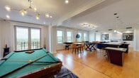 This expansive single-floor space blends refined architectural detailing with contemporary furnishings across several well-defined living and entertainment areas. Throughout, warm hardwood flooring, crown molding, recessed lighting, and decorative column accents establish a cohesive and polished aesthetic.

The kitchen is a standout, featuring sleek white cabinetry, a subway-tile backsplash, professional-grade stainless appliances, and a generous island topped with a light stone surface. A striking row of sculptural black pendant lights anchors the space, while emerald green velvet seating adds a bold design note.

An adjoining dining and lounge area includes a wall-mounted television and a mirrored wet bar with glass shelving, creating an ideal entertaining hub. The living room offers a refined sitting area centered around a black marble fireplace with a classic white mantel, complemented by a tufted navy ottoman and a velvet sofa. A secondary lounge features blue velvet armchairs, a live-edge coffee table, and panoramic windows with expansive views.

The dedicated game room accommodates a full-size pool table, a shuffleboard table, and casual seating, all bathed in natural light from large double-hung windows that frame scenic outdoor vistas. A private balcony further enhances the connection to the surrounding landscape.