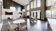 Located in Scottsdale, Arizona, this thoughtfully designed communal space delivers a polished, contemporary aesthetic across a single open floor plan. Soaring double-height ceilings create an impressive sense of volume, while floor-to-ceiling windows and glass French doors flood the interior with natural light and offer views of the surrounding area.

The kitchen is a standout feature, centered around a large white quartz island with bar seating and dramatic clustered pendant lighting overhead. Stainless steel appliances line the back wall, which is finished with a textured mosaic tile backsplash that adds warmth and visual interest. Dark hardwood flooring grounds the space with a rich contrast to the bright cabinetry and countertops.

The lounge area is divided into distinct seating zones. One area features a dark upholstered sofa and chairs arranged around a media wall clad in a combination of wood paneling and light stone tile, anchored by a wall-mounted flat-screen television. A separate conversational nook includes white accent chairs and a marble-top coffee table set atop a geometric patterned area rug. Dining tables with upholstered chairs are positioned near the windows, providing casual gathering spots.

Throughout, the design balances warm and cool tones with a curated mix of textures, creating an inviting yet sophisticated environment well-suited for socializing and gathering.