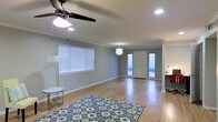 This single-story space offers an open, airy layout with warm hardwood flooring that extends throughout the main living area. The room is finished with clean crown molding and neutral gray walls, creating a modern yet inviting atmosphere. Recessed lighting is complemented by a ceiling fan with integrated light and a decorative pendant fixture, providing versatile illumination options. A wide window fitted with horizontal blinds allows ample natural light to fill the space, further enhanced by sidelight panels flanking the front entry door. The entryway is framed by a classic paneled door, contributing to a tidy, well-defined transition between the interior and exterior. The open floor plan connects the living area seamlessly to an adjacent workspace or desk area, visible along the right side of the room. The overall ambiance is clean and contemporary, with thoughtful architectural details such as the crown molding and consistent flooring creating a cohesive, polished feel throughout. The generous proportions of the main living area provide flexibility for a variety of furniture arrangements and functional uses.