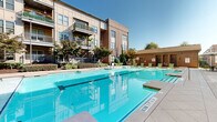 Located at 3000 Forrest Walk in Roswell, GA, this single-floor communal living space serves as a sophisticated outdoor lounge and gathering area within a modern residential community. The space is defined by its striking architectural character, blending warm natural stone brick walls with elegant arched openings, exposed wood-beam ceilings, and geometric stone-tile flooring that lends a refined yet welcoming atmosphere.

The living room area is thoughtfully appointed with plush, neutral-toned seating, accent chairs in bold orange, and sleek coffee tables, all arranged beneath ceiling fans that ensure year-round comfort. A wall-mounted flat-screen TV anchors the lounge, while floor-to-ceiling black-framed glass walls provide a seamless visual connection to the interior clubhouse beyond, flooding the space with natural light.

Layered staircases with iron railings and stone steps add architectural depth and connect to multiple outdoor levels, leading out to the broader community grounds. Adjacent to the lounge, a expansive resort-style swimming pool is framed by lounge seating and lush landscaping, set against the backdrop of the multi-story residential building. This space masterfully combines indoor comfort with outdoor living, creating a vibrant, upscale amenity area ideal for relaxation and socializing.