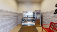 This dedicated pet grooming space is thoughtfully designed to provide a clean, functional environment for pet care. The room is finished with durable, large-format stone-look ceramic tiles that run halfway up the walls, creating a practical and attractive wainscoting effect, while the upper walls are finished in neutral tones. The light beige tile flooring is easy to maintain and complements the overall aesthetic.

The centerpiece of the space is a stainless steel Evolution Dog Wash self-service station, complete with an anti-fatigue rubber mat in front for user comfort. The unit features an integrated payment system, a splash guard enclosure, and a hose attachment, offering a hygienic and convenient bathing solution. Adjacent to the wash station, a vibrant red decorative bench provides seating for pet owners, accompanied by wall-mounted leash hooks and pet-themed décor that adds a welcoming character to the room.

A wall-mounted mini-split air conditioning unit ensures year-round climate control, maintaining a comfortable environment for both pets and owners. A windowed entry door allows natural light into the space, while a waste bin and fire safety equipment are conveniently positioned near the entrance, underscoring the room's practical, well-organized design.
