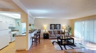 This single-floor residence offers a thoughtfully designed open-concept layout that seamlessly connects the kitchen, dining, and living areas into one cohesive and inviting space. The warm-toned hardwood flooring extends throughout, lending a sense of continuity and warmth to the entire floor plan.

The kitchen features light wood cabinetry and a raised breakfast bar with seating for two, providing both a casual dining option and a natural dividing element between the cooking and living spaces. A window above the kitchen counter allows natural light to filter in, brightening the workspace.

The living area is generously proportioned, centered around a comfortable sectional sofa and a dark-framed coffee table set atop a stylish area rug. Matching table lamps cast a warm, ambient glow, while a pair of framed wall art pieces add a decorative focal point. A lush indoor plant in the corner brings a touch of natural greenery to the space.

The textured ceiling adds subtle visual interest overhead, while the neutral, warm wall tones create a calm and versatile backdrop throughout. The overall ambiance is cozy yet functional, making this residence well-suited for both everyday living and casual entertaining.
