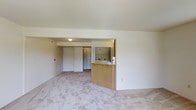 This single-floor unit offers a clean, functional layout designed for comfortable everyday living. The open-concept living area is finished with plush wall-to-wall carpeting in a neutral tone, creating a warm and inviting atmosphere throughout the space. A defining feature of the layout is the kitchen peninsula, crafted with light wood cabinetry and a built-in sink, which seamlessly connects the kitchen to the main living area. A large mirror above the peninsula enhances the sense of openness and reflects natural light into the room. The entryway is clearly defined, featuring a solid white door that leads into the unit with ease. The interior is painted in soft, neutral tones from floor to ceiling, lending a bright and airy feel to the entire space. Flush-mounted ceiling lighting provides consistent illumination throughout. The straightforward, uncluttered design of the unit makes it highly adaptable for a variety of furnishing arrangements and personal styles. Overall, the space strikes a balance between practicality and comfort, with a cohesive design language that emphasizes clean lines and an open, welcoming ambiance.