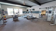 Located at 3800 14th Ave SE in Lacey, Washington, this fitness center is a well-appointed, single-floor facility designed to support a comprehensive workout experience. The space is thoughtfully divided into distinct exercise zones, each catering to different training needs.

The cardio area is equipped with multiple treadmills, an elliptical trainer, and a stationary bike, all positioned to take advantage of natural light streaming through windows that offer pleasant outdoor views of surrounding greenery. Wall-mounted flat-screen televisions are conveniently positioned above the cardio equipment, allowing users to stay entertained during their workouts.

The strength training zone features a multi-station cable machine, a rack of dumbbells, and a weight bench, providing a full range of resistance training options. Full-length mirrors line the walls in this area, enhancing the sense of space and allowing users to monitor their form. A medicine ball rack near the mirrored wall adds additional versatility for functional training.

The interior design features a neutral color palette with a distinctive teal accent stripe running along the walls, complemented by dark trim details near the ceiling that add a modern, polished character. Textured ceilings and ceiling fans contribute to the overall comfort of the space. French-style doors connect the fitness room to an adjacent hallway, ensuring easy accessibility throughout the facility.