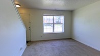 This two-story residence offers a practical and straightforward layout across two levels. The kitchen is equipped with warm-toned wooden cabinetry providing generous storage space, a double-basin sink, a white dishwasher, and a white electric range. A checkered tile floor adds a classic touch, and a fluorescent ceiling fixture ensures well-lit meal preparation. A hallway visible from the kitchen leads toward a staircase connecting to the upper floor, suggesting a logical and accessible flow between spaces.

The living area features neutral-toned carpeting that lends a comfortable, understated feel to the room. A large double-hung window allows natural light to fill the space and offers views of the surrounding exterior. The textured ceiling is consistent throughout the home, contributing to a uniform aesthetic. The front entry door is conveniently located within the living area, providing direct access to the outdoors. Overall, the space presents a clean, functional environment with practical finishes suited for everyday living.