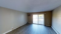 This single-floor space offers a straightforward and functional layout designed for comfortable everyday living. The main living area is finished with smooth, hardwood-style laminate flooring that runs throughout, providing a clean and cohesive look underfoot. A textured popcorn ceiling adds a classic touch overhead, while neutral beige walls create a versatile backdrop suitable for a variety of furnishing styles.

Natural light is a notable feature of the living area, courtesy of a large sliding glass door that opens onto an outdoor balcony. The balcony offers views of surrounding greenery, bringing a sense of the outdoors into the interior. The sliding door is framed with white trim and complemented by a privacy curtain panel.

Climate control is handled by a wall-mounted air conditioning unit positioned near the sliding door, supplemented by a baseboard heater running along the exterior wall — ensuring year-round comfort. Electrical outlets are conveniently placed along the walls to support various furniture arrangements.

Overall, the space presents a clean, move-in-ready environment with practical amenities and a simple, uncluttered aesthetic that makes it easy to personalize and adapt to individual needs.