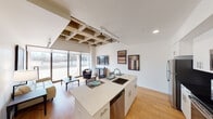 This contemporary single-floor space blends industrial design elements with modern finishes to create a stylish and functional living environment. The open-concept layout seamlessly connects the kitchen and living area, fostering a sense of openness and flow throughout.

The kitchen is a standout feature, anchored by a generous island with a light quartz countertop, an undermount sink, and bar seating for casual dining. On the opposite side, tall white cabinetry pairs with dark granite countertops, a black tile backsplash, and a full suite of stainless steel appliances — including a refrigerator, range, microwave, and dishwasher — delivering both style and practicality.

An eye-catching exposed wood beam grid detail on the ceiling above the kitchen and dining area adds a warm, industrial character to the space. Track lighting runs alongside it, providing focused illumination throughout. The living area benefits from abundant natural light streaming through large sliding glass doors that open to an outdoor area, offering pleasant exterior views. Warm hardwood flooring runs throughout, tying the spaces together cohesively.

The bedroom is a quiet, minimalist retreat with neutral walls and the continuation of the exposed ceiling detail. Clean white walls and thoughtful layout throughout the unit create a bright, airy ambiance well-suited for modern living.