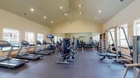 This fitness center is a comprehensively outfitted facility designed to support a wide range of workout routines. The main exercise area is defined by a dramatic vaulted ceiling that rises to an impressive height, creating an open, airy atmosphere enhanced by recessed lighting and a large wall-to-wall mirror that visually expands the space. Durable rubber flooring throughout provides both safety and comfort underfoot.

The cardio zone features multiple treadmills, elliptical trainers, and upright stationary bikes, all positioned near large windows that offer natural light and views of the surrounding outdoor area. Wall-mounted televisions are conveniently placed for entertainment during workouts. The strength training section is well-stocked with a full dumbbell rack, cable machines, a functional trainer, and a variety of selectorized weight machines and benches to accommodate diverse training needs.

A dedicated functional training alcove offers medicine balls, stability balls, foam rollers, and a large flat-screen display with an interactive fitness platform, enabling guided workout sessions. Additional equipment such as yoga mats is also available in this zone. The facility flows between its sections through open doorways, maintaining a cohesive and spacious feel throughout. Tasteful wall art, including a prominent yin-yang canvas, adds character to the overall environment.