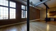 This 2-bedroom, 2-bathroom residence offers a generous single-floor layout with a distinctive industrial-loft aesthetic. The open living area serves as the centerpiece of the space, featuring warm hardwood floors that complement the exposed red brick walls, which add texture and character throughout. Large multi-pane windows line the exterior walls, allowing abundant natural light to pour in while offering views of the surrounding urban environment.

Overhead, exposed metal ductwork and structural columns contribute to the space's industrial character, lending an authentic, converted-building feel. The high ceilings enhance the sense of openness and airiness in the main living area, making it feel particularly expansive and versatile for various furniture arrangements and uses.

The layout accommodates two full bedrooms and two bathrooms, providing comfortable and functional private spaces alongside the open communal areas. The combination of raw industrial materials—brick, steel, and exposed mechanical systems—balanced with the warmth of hardwood flooring creates a dynamic contrast that defines the overall ambiance of this thoughtfully designed urban residence.