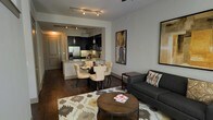 This single-story Dallas residence showcases a thoughtfully designed open-concept layout that seamlessly connects the living, dining, and kitchen areas into one cohesive space. Rich, dark hardwood flooring runs throughout, grounding the interior with warmth and continuity.

The kitchen is outfitted with sleek dark cabinetry, stainless steel appliances including a French door refrigerator, and a light-toned countertop peninsula that doubles as a casual dining surface. Pendant lighting suspended above the peninsula adds a refined touch, complementing the track lighting overhead.

The dining area sits just adjacent to the kitchen, accommodating a four-person table with elegant upholstered chairs, making it ideal for both everyday meals and entertaining. Transitioning naturally into the living area, the space features a charcoal sofa accented with decorative pillows, a rustic round wooden coffee table, and a geometric-patterned area rug that anchors the seating arrangement. Framed artwork on the walls adds character and visual interest. A ceiling fan overhead provides both comfort and function. The overall ambiance is modern and well-appointed, balancing contemporary design with inviting warmth.