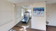 This single-floor fitness room offers a practical and functional space designed to support cardiovascular exercise and general wellness. The room is outfitted with Precor equipment, including a treadmill and an upright stationary bike, positioned to take full advantage of the natural light streaming in through multiple windows fitted with adjustable blinds. The windows provide pleasant views of the surrounding outdoor environment, contributing to an airy and motivating atmosphere during workouts.

Large wall-to-ceiling mirrors are strategically placed to visually expand the room and allow users to monitor their form while exercising. The mirrors also reflect the natural light, brightening the interior further. The flooring consists of durable, low-pile carpet well-suited for a fitness setting, while a suspended acoustic tile ceiling with recessed fluorescent lighting ensures consistent illumination throughout.

The space also includes a small entryway area with a folding chair, providing a spot to rest or change footwear before or after a session. Tasteful framed artwork and a colorful painted canvas depicting a nautical scene decorate the walls, adding a touch of personality to the utilitarian space. A security camera and smoke detector are also present, reflecting attention to safety. Overall, this fitness room is a clean, bright, and thoughtfully arranged space suitable for daily cardio workouts.