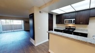 This single-floor residence offers a thoughtfully designed layout that balances functionality and style. The kitchen is a standout feature, outfitted with rich, dark wood cabinetry that provides ample storage, granite countertops with subtle veining, and stainless steel appliances including a gas range and built-in microwave. A skylight panel overhead floods the kitchen with natural light, creating a bright and inviting workspace. The kitchen opens to the rest of the living space via a peninsula counter, promoting an open, connected feel throughout the floor plan. Durable hardwood-style flooring extends seamlessly from the kitchen through the hallway and into the living area, lending a warm and cohesive aesthetic. The living area is visible in the background, benefiting from additional natural light through a sliding glass door or window. Neutral, warm-toned walls complement both the dark cabinetry and the flooring, creating a balanced interior palette. Practical details such as HVAC vents, a wall-mounted AC unit, and ample closet access add to the everyday livability of this well-maintained space.