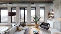 This contemporary urban residence masterfully blends industrial and modern design across a single thoughtfully laid-out floor. Exposed concrete ceilings with visible ductwork lend an authentic loft aesthetic throughout, while light hardwood flooring and crisp white walls create a clean, sophisticated contrast.

The open living area is a standout, featuring a sleek built-in linear fireplace with a floating wood mantle and curated gallery wall. Oversized black-framed windows frame sweeping, high-rise city skyline views, flooding the space with natural light. A separate dining area accommodates a full glass dining table beneath the same dramatic city panorama.

The kitchen is outfitted with high-gloss white cabinetry reaching the ceiling, stainless steel appliances including a French door refrigerator, gas cooktop, and dishwasher, all set against a clean tile backsplash and stone countertops.

The bedrooms offer a serene retreat, each with warm hardwood floors and the signature exposed concrete ceiling. One bedroom features a spacious walk-in closet with built-in shelving and double hanging rods. Bathrooms are finished with backlit LED mirrors, undermount sinks, and wood-toned vanities. A dedicated laundry room houses a full-size Whirlpool washer and dryer with overhead hanging storage.