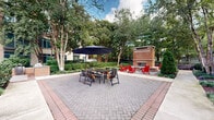 Located at 2121 N Westmoreland Street in Arlington, Virginia, this well-appointed outdoor courtyard serves as a central gathering space within a multi-story brick residential complex. The courtyard is anchored by a striking standalone brick fireplace, surrounded by vibrant red Adirondack chairs that invite relaxation and socializing. A spacious herringbone-patterned brick patio provides ample room for outdoor dining, complete with a large table, chairs, and a navy blue market umbrella offering shade. The space is enclosed on multiple sides by the building's classic brick facade, featuring large windows and balconies that overlook the courtyard below. Mature deciduous and evergreen trees rise throughout the space, providing natural canopy and a sense of seclusion. Thoughtfully designed landscaping includes neatly trimmed hedges, ornamental shrubs, ground cover plantings, and mulched garden beds that add texture and greenery throughout. A concrete walkway winds through the garden beds, connecting different areas of the courtyard and leading toward additional seating. The overall ambiance is one of a tranquil, well-maintained urban retreat that balances natural elements with functional outdoor amenities, offering residents a welcoming environment for both quiet relaxation and communal gatherings.
