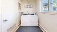 Located at 2150 W 15th St in Loveland, Colorado, this shared laundry room is a compact, efficiently arranged space designed to serve its users with ease. The room features light-toned luxury vinyl plank flooring that contributes to a clean, bright atmosphere throughout. At the heart of the space sit two commercial-grade Speed Queen machines — a top-load washer and a front-load dryer — both coin-operated and well-maintained for shared use. A utility shelf above the machines provides a convenient surface for laundry supplies. The walls are finished in a neutral off-white tone, keeping the space feeling open and tidy. A window on one wall allows natural light to filter in, offering a view of the surrounding greenery. Baseboard heaters line the perimeter, ensuring the room remains comfortable year-round. A cork bulletin board displays notices and building information, while framed instructional signage above the machines keeps users informed of proper operating procedures. A wall clock and a security camera round out the functional amenities of this well-organized laundry facility, making it a practical and straightforward shared resource for residents.