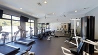 This fitness center is designed to provide a comprehensive workout environment within a single, open-plan floor. The space is outfitted with a wide variety of exercise equipment, including multiple treadmills, elliptical trainers, and weight benches, catering to both cardio and strength training needs. Full-length mirrored walls line one side of the room, visually expanding the space and allowing users to monitor their form during workouts. Large floor-to-ceiling windows along the opposite wall flood the interior with natural light and offer views of an outdoor area, creating an open and energizing atmosphere. The flooring consists of durable black rubber matting, designed to withstand heavy equipment and high-traffic use. Overhead, multiple ceiling fans are evenly distributed across the white ceiling, supplemented by recessed lighting to ensure a consistently well-lit environment. A wall clock is also present, helping users manage their workout sessions. The overall ambiance is clean, functional, and modern, making this fitness center a practical and well-appointed space for a full range of exercise activities.
