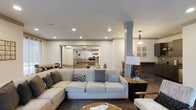 This single-floor space offers an open-concept layout that connects multiple living areas in a cohesive and functional design. The main living area is anchored by a generously sized sectional sofa and complemented by a stylish round coffee table, creating an inviting centerpiece for relaxation and social gatherings. Recessed lighting in the white ceiling provides consistent, ambient illumination throughout the space, while a decorative pendant light adds a refined accent near the fireplace.

A prominent structural column with a clean, grey finish subtly defines the transition between zones without interrupting the open flow. Adjacent to the main seating area, a fireplace with a sleek surround serves as a striking focal point, flanked by curated wall art and a statement table lamp. The warm hardwood flooring ties the spaces together, lending a natural, grounded quality to the interior.

Looking beyond the main living space, additional furnished seating areas are visible, highlighting the depth and scale of the layout. Neutral tones of white, grey, and beige are used consistently throughout, creating a calm and sophisticated atmosphere. The thoughtful combination of modern furnishings, layered lighting, and architectural details makes this a versatile and welcoming environment.